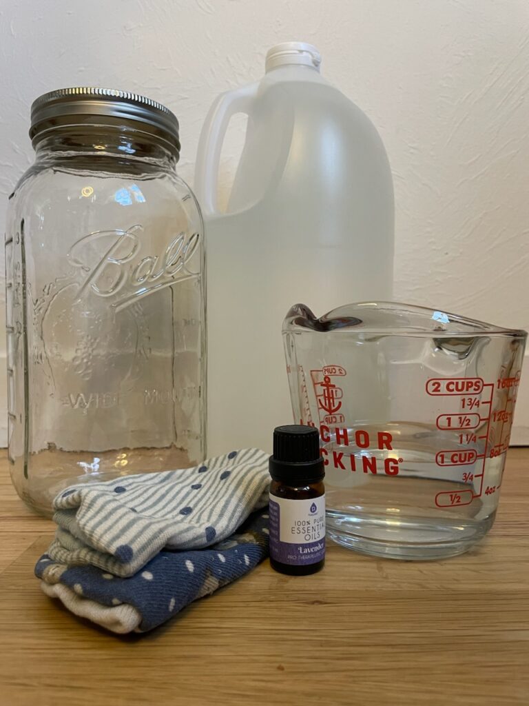 a glass mason jar, a jug of vinegar, a measuring cup filled halfway with liquid, lavender essential oil, and two wash cloths all sitting next to each other. 