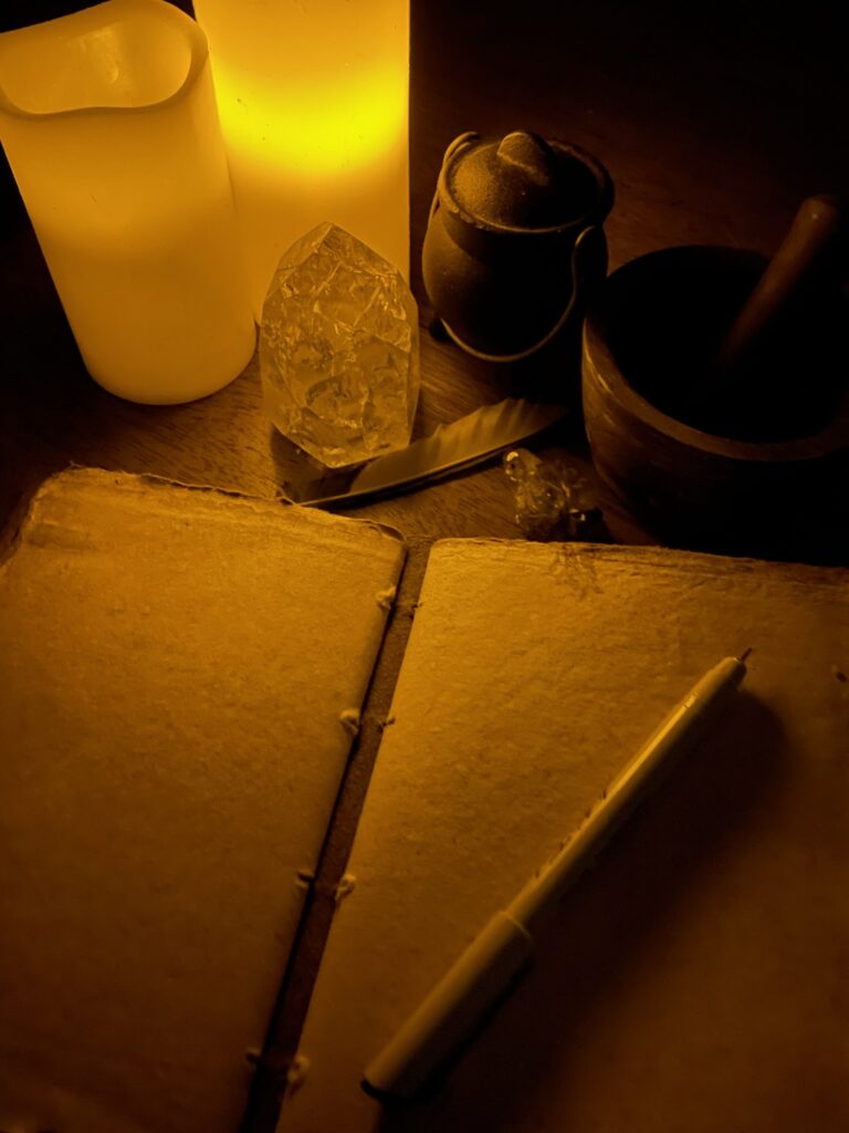 a blank book with a pen on top of its open pages, with a pestle and mortar, a small cauldron, a large and small crystal, and a feather all above the book sitting in candle light.