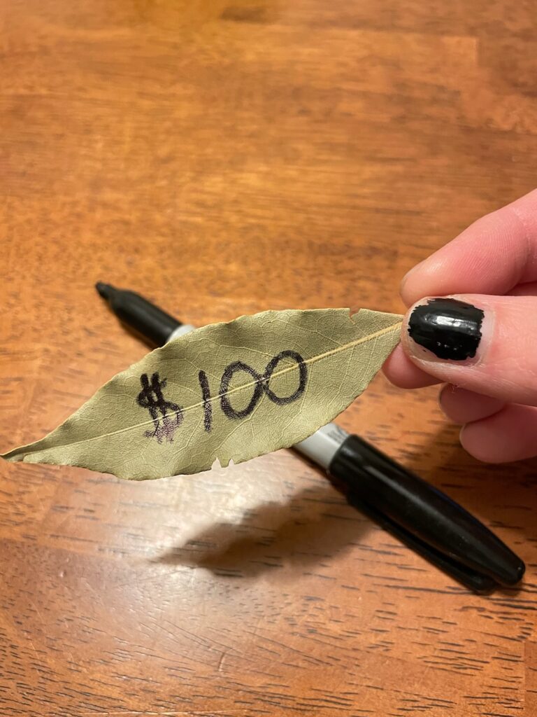 a hand with black nail polish on the thumb finger, holding a bay leaf that has $100 written on it and a black open marker laying on the table. 